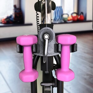 May include: Two pink dumbbells are attached to a black exercise bike. The dumbbells are held in place by a black holder.