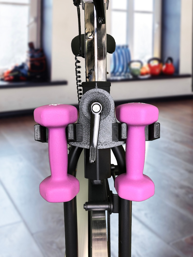 May include: Two pink dumbbells are attached to a black exercise bike. The dumbbells are held in place by a black holder.