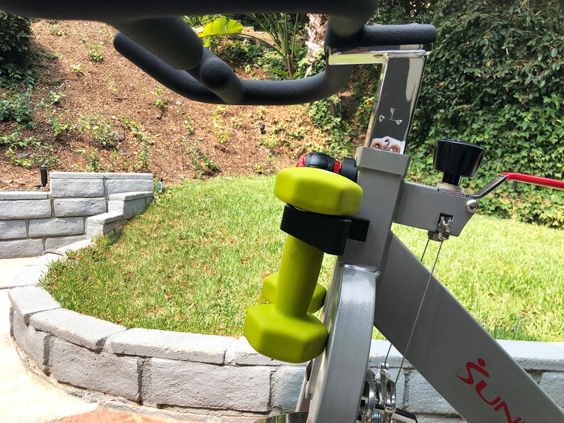 May include: A gray exercise bike with a black water bottle holder and a pair of green dumbbells attached to the handlebars. The bike has a red "Sunny" logo on the side.