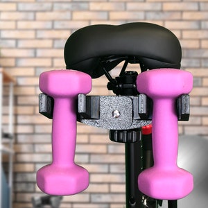 May include: A black exercise bike seat with two pink dumbbells attached to a black metal holder.