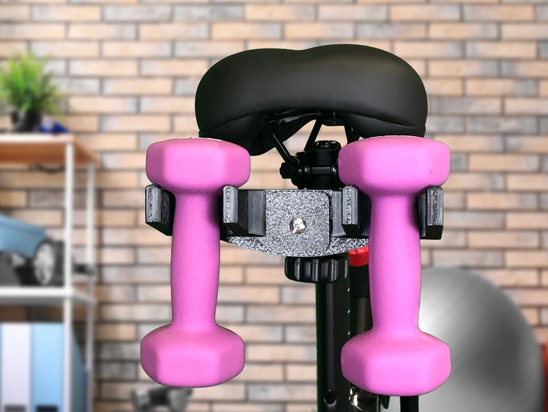 May include: A black exercise bike seat with two pink dumbbells attached to a black metal holder.