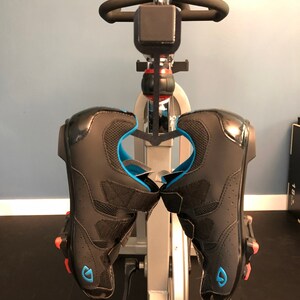 May include: A pair of black cycling shoes with blue and red accents are hanging on a stationary bike.