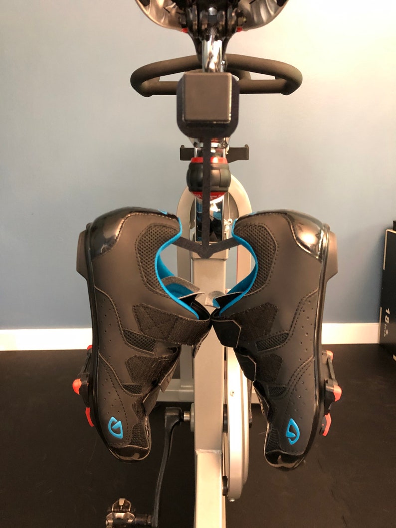 May include: A pair of black cycling shoes with blue and red accents are hanging on a stationary bike.