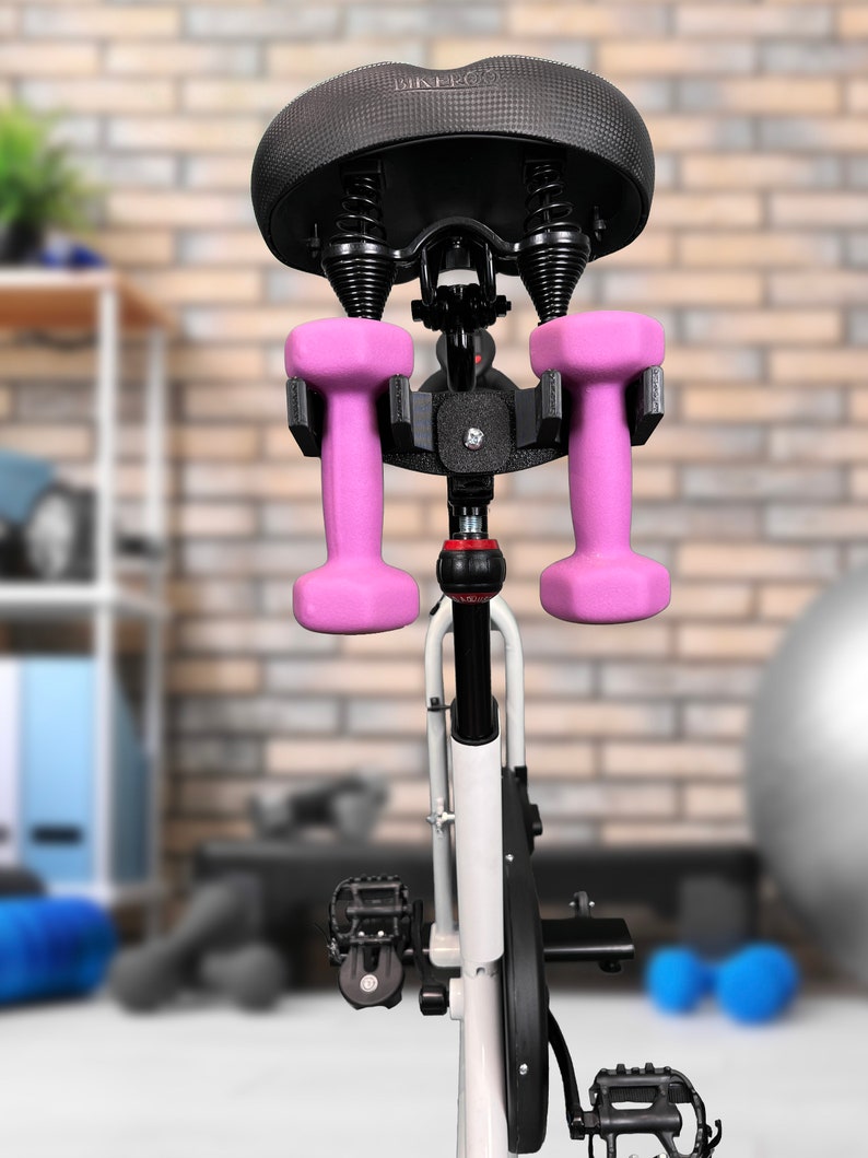 May include: A black exercise bike with a padded seat and a holder for two pink dumbbells attached to the seat post.