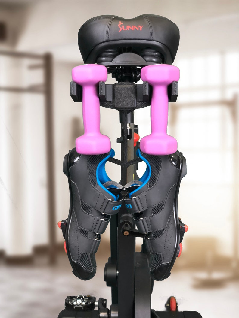 Pu&ograve; includere: Bicicletta da esercizio nera con una sella nera, manubri rosa e scarpe da ciclismo nere appese alla bicicletta. La bicicletta ha un telaio nero e una base argentata. La sella ha la scritta "SUNNY" stampata in rosso.