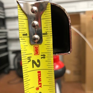 May include: A close-up of a yellow Stanley measuring tape showing a measurement of approximately 2 inches. The tape is positioned against a black, curved metal object. The background is blurred, with hints of a garage setting.