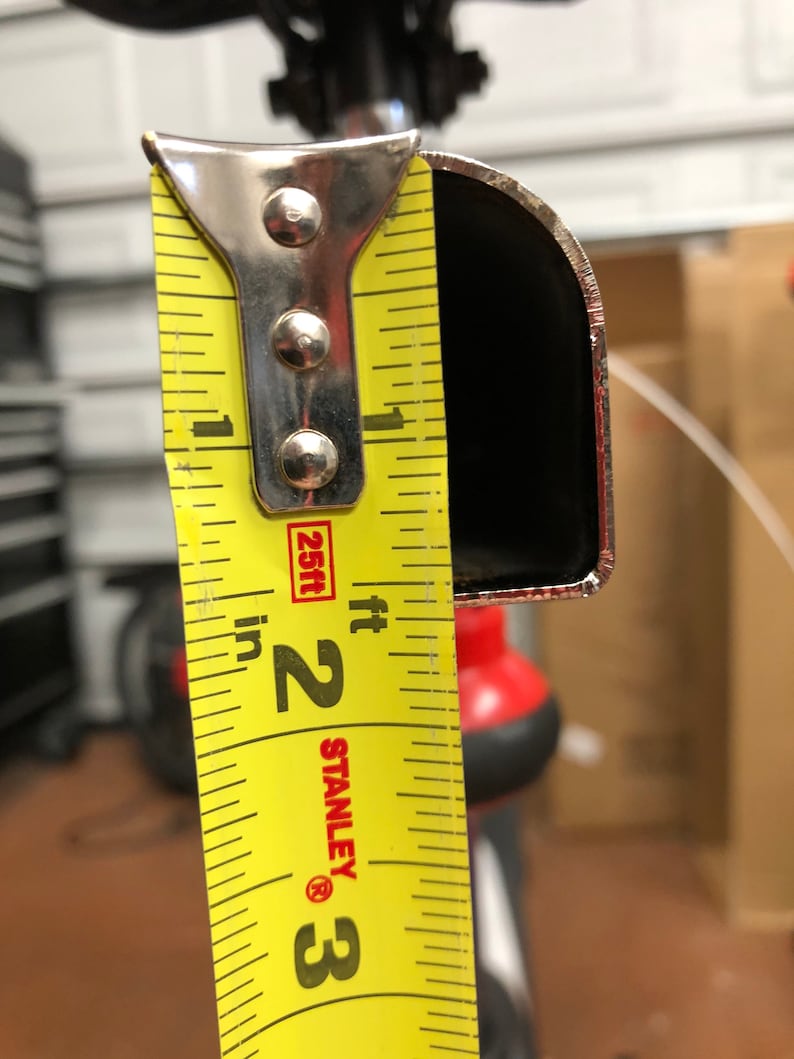 May include: A close-up of a yellow Stanley measuring tape showing a measurement of approximately 2 inches. The tape is positioned against a black, curved metal object. The background is blurred, with hints of a garage setting.