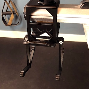 May include: Black metal exercise equipment attachment with a square top and adjustable legs. The attachment has a knob on top and is designed to clamp onto a bar. A pair of exercise straps hangs in the background.