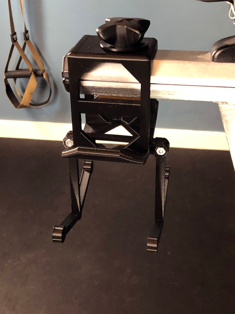 May include: Black metal exercise equipment attachment with a square top and adjustable legs. The attachment has a knob on top and is designed to clamp onto a bar. A pair of exercise straps hangs in the background.