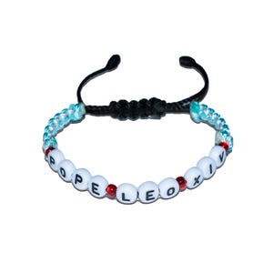 May include: Adjustable bracelet with white letter beads spelling "POPE LEO XIV". The bracelet features red and blue accents, with a black adjustable cord. A unique accessory.