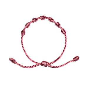 May include: A maroon-colored bracelet featuring a series of woven knots. The adjustable bracelet is made of a cord material and has a sliding knot closure. The bracelet's design includes seven decorative knots along the main band, creating a unique and stylish accessory.