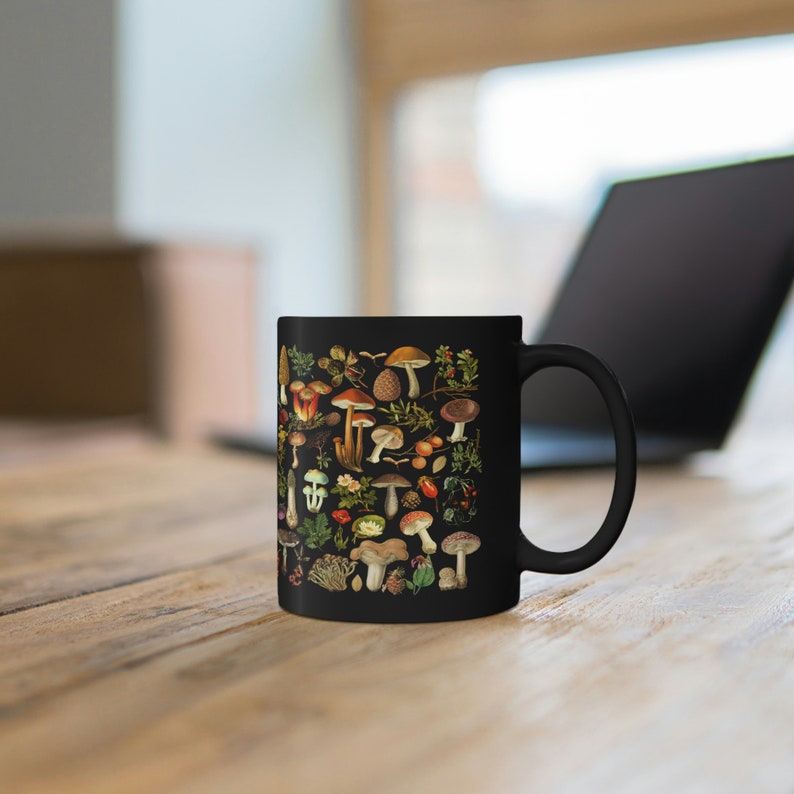 May include: A black ceramic mug with a colorful illustration of various mushrooms and foliage. The mug has a white handle.