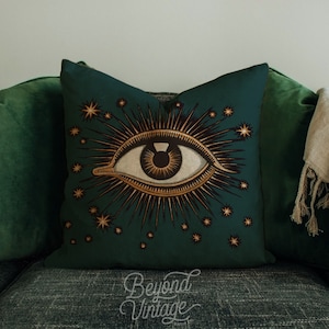 Könnte beinhalten: Ein grünes Kissen mit einem goldenen Augen- und Sternendesign. Das Auge ist von einem goldenen Sonnenstrahl und Sternen umgeben.