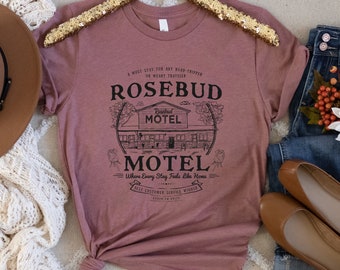 May include: A mauve t-shirt featuring a black graphic design of a motel called "Rosebud Motel". The graphic includes the text "Where Every Stay Feels Like Home" and "Best Customer Service Winner".