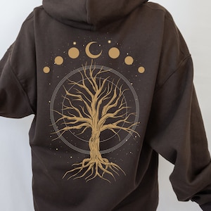 May include: A dark brown hoodie featuring a gold-colored tree design with a crescent moon and celestial elements. The tree has detailed branches and roots, set within a circular frame. The hoodie has a hood and long sleeves.