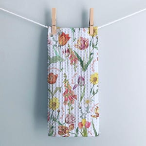 Op de afbeelding: Een witte wafeldoek met een bloemenpatroon van tulpen, vingerhoedenkruid en andere bloemen in rood, oranje, geel en paars. De handdoek hangt aan een waslijn met houten wasknijpers.
