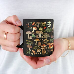May include: A black ceramic mug with a colorful illustration of various mushrooms and leaves. The mug has a handle on the left side.