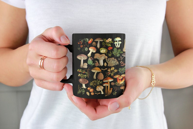 May include: A black ceramic mug with a colorful illustration of various mushrooms and leaves. The mug has a handle on the left side.