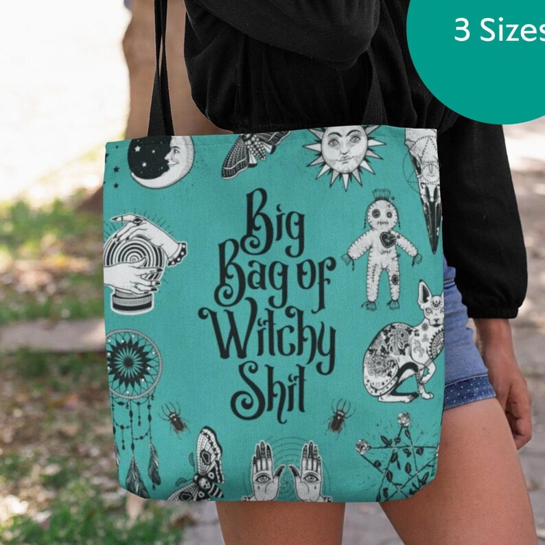 A person is holding a teal-colored tote bag with various whimsical and mystical illustrations, including a moon, sun, cat, and other fantastical elements.