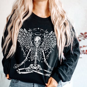 May include: Black sweatshirt with a white graphic of a skeleton with angel wings, a halo, and a meditating pose. The design includes stars and lines. The sweatshirt has long sleeves and a crew neck.