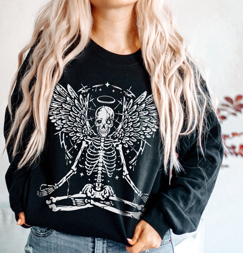 May include: Black sweatshirt with a white graphic of a skeleton with angel wings, a halo, and a meditating pose. The design includes stars and lines. The sweatshirt has long sleeves and a crew neck.