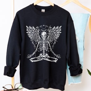 May include: Black sweatshirt with a white graphic of a meditating skeleton with angel wings and a halo. The design includes stars and a circular pattern. The sweatshirt is displayed on a wooden hanger, with a pair of jeans visible.