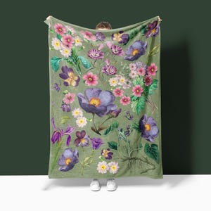 May include: A green throw blanket with a floral print featuring purple, pink, and white flowers. The blanket is made of soft, plush fabric and is perfect for adding a touch of nature to your home.