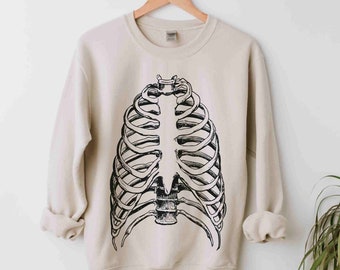 Op de afbeelding: Een beige sweatshirt met een zwart-witte illustratie van een menselijke ribbenkast. De afbeelding staat in het midden op de voorkant van het kledingstuk met lange mouwen. Het sweatshirt hangt aan een houten kleerhanger tegen een witte achtergrond.