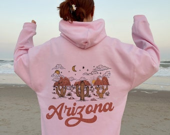 Può includere: Felpa rosa con una grafica di un paesaggio desertico con cactus e la parola "Arizona" in un font vintage.