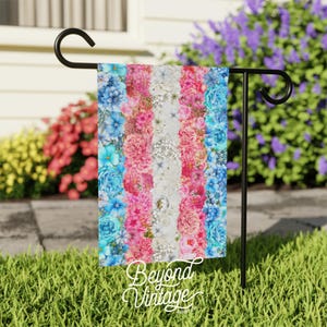 May include: A garden flag with a transgender pride flag design. The flag is made of fabric and features a pattern of blue, pink, and white flowers. The text "Beyond Vintage" is printed at the bottom of the flag.