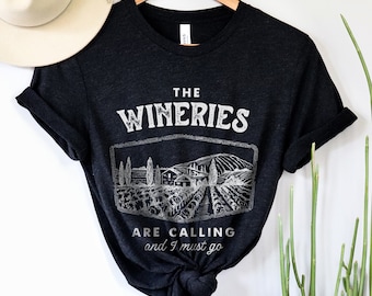 May include: Black t-shirt with a white graphic design that says "The Wineries Are Calling and I Must Go". The graphic features a vineyard scene with a house and trees.