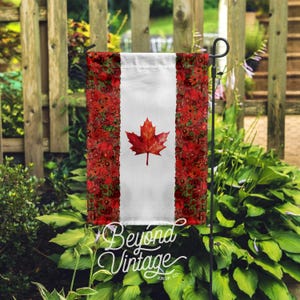 Puede incluir: Una bandera de jardín con el diseño de la bandera canadiense. La bandera tiene un centro blanco con una hoja de arce roja y bordes florales rojos. Las palabras "Beyond Vintage Shop Co." están escritas en blanco en la parte inferior.