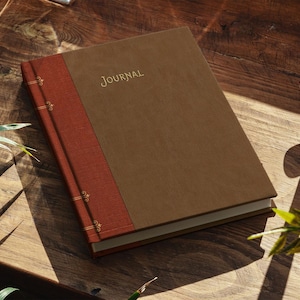May include: A rectangular journal with a brown cover and a textured rust-colored spine. The word JOURNAL is embossed in gold on the front cover. The spine features gold decorative lines and small floral accents along the vertical edge.