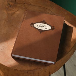 A brown hardcover book titled "Book of Curses" rests on a wooden stool against a green wall.