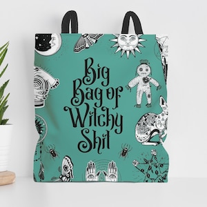 A green tote bag with a whimsical, cartoonish design featuring various mystical and supernatural elements, such as a moon, sun, spider, and voodoo doll. The bag has a playful, humorous text that reads "Big Bag of Witchy Shit" and is displayed on a shelf next to a potted plant.