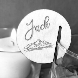 K&ouml;nnte beinhalten: Eine Holz-Platzkarte mit dem Namen "Jack" in kursiv geschriebener Schrift. Die Karte zeigt ein Bergpanorama und einen Schlitz zum Halten einer Karte.
