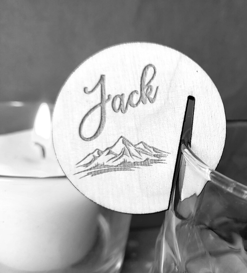 K&ouml;nnte beinhalten: Eine Holz-Platzkarte mit dem Namen "Jack" in kursiv geschriebener Schrift. Die Karte zeigt ein Bergpanorama und einen Schlitz zum Halten einer Karte.