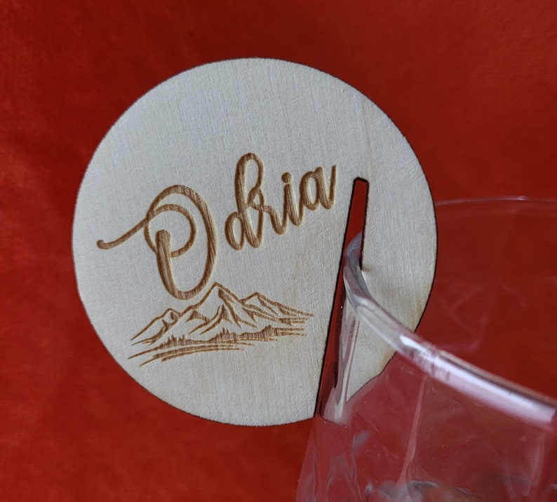 K&ouml;nnte beinhalten: Ein Holz-Getr&auml;nke-Marker mit dem Namen "Olivia" eingraviert. Der Marker hat ein Bergketten-Design und einen Ausschnitt f&uuml;r ein Glas.