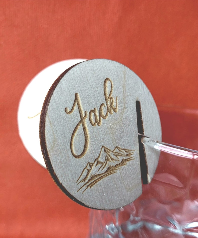 K&ouml;nnte beinhalten: Ein Holz-Getr&auml;nke-Marker mit dem Namen "Jack" eingraviert. Der Marker zeigt auch ein Bergpanorama.