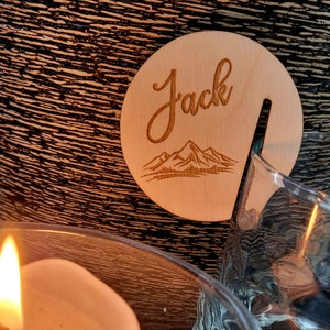 K&ouml;nnte beinhalten: Ein Holz-Untersetzer mit dem eingravierten Namen "Jack". Der Untersetzer zeigt ein Bergpanorama und eine Aussparung f&uuml;r ein Glas.