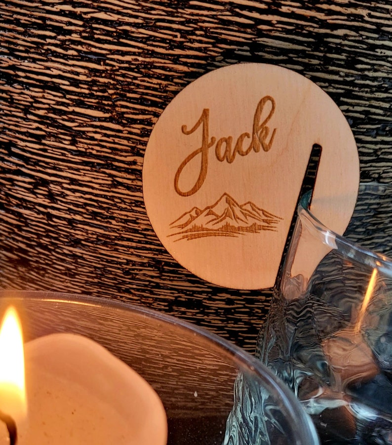 K&ouml;nnte beinhalten: Ein Holz-Untersetzer mit dem eingravierten Namen "Jack". Der Untersetzer zeigt ein Bergpanorama und eine Aussparung f&uuml;r ein Glas.