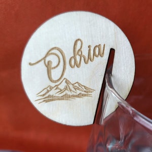 K&ouml;nnte beinhalten: Ein Holz-Getr&auml;nke-Marker mit dem Namen "Olivia" eingraviert. Der Marker hat ein Bergpanorama-Design und einen Schlitz zum Befestigen an einem Glas.
