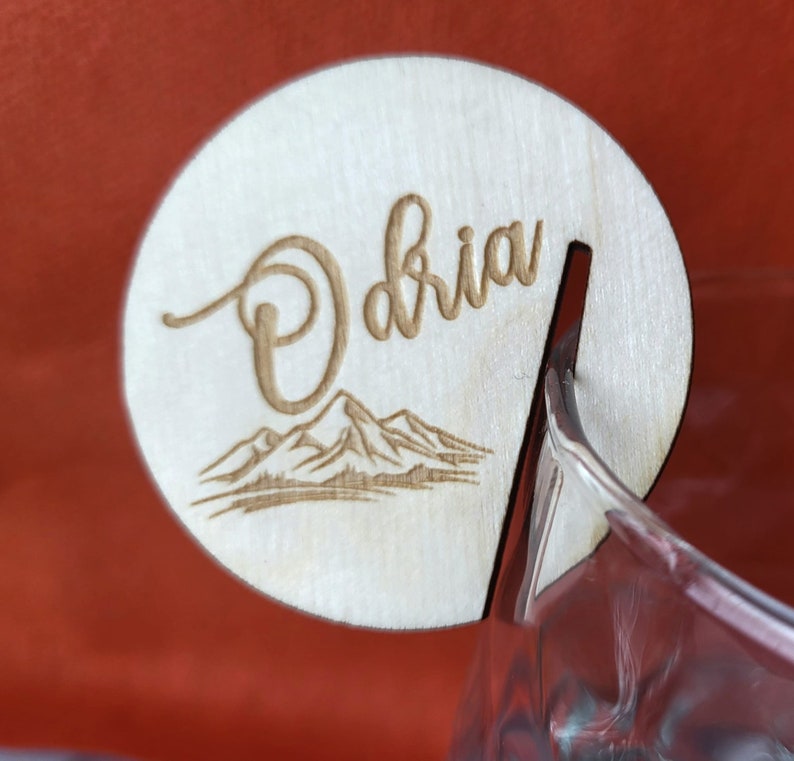 K&ouml;nnte beinhalten: Ein Holz-Getr&auml;nke-Marker mit dem Namen "Olivia" eingraviert. Der Marker hat ein Bergpanorama-Design und einen Schlitz zum Befestigen an einem Glas.