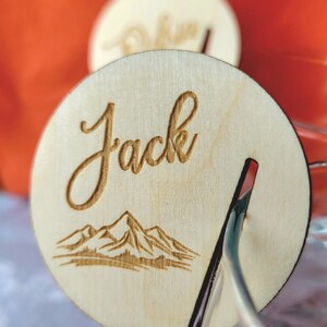 K&ouml;nnte beinhalten: Eine Holz-Platzkarte mit dem Namen "Jack" in kursiv geschriebener Schrift. Die Karte zeigt eine Illustration einer Bergkette und einen Schlitz f&uuml;r einen Halter.