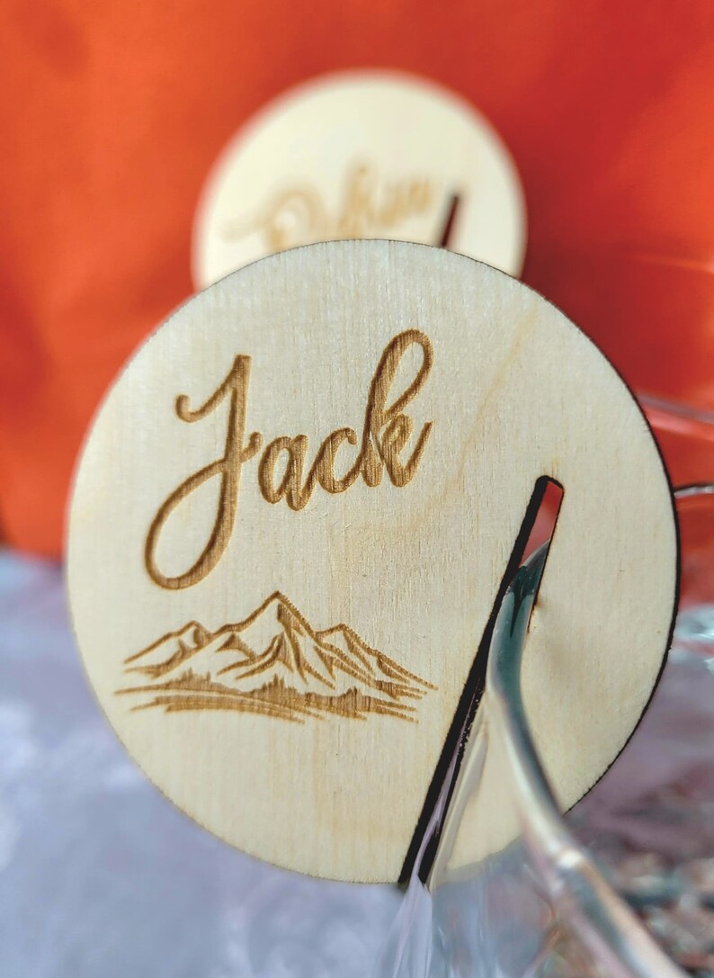 K&ouml;nnte beinhalten: Eine Holz-Platzkarte mit dem Namen "Jack" in kursiv geschriebener Schrift. Die Karte zeigt eine Illustration einer Bergkette und einen Schlitz f&uuml;r einen Halter.