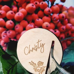 May include: A round wooden place card with the name "Christy" engraved in cursive script. The place card has a small floral design engraved in the bottom right corner.