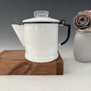 以下が含まれることがあります： 透明なガラスの蓋と黒いハンドルが付いた白いエナメルコーヒーポット。コーヒーポットは木製の表面に置かれています。注ぎ口はポットの左側にあります。