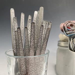 Puede incluir: Un tarro de vidrio lleno de un conjunto de cubiertos plateados con un diseño floral adornado. Los cubiertos están dispuestos verticalmente en el tarro.