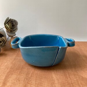 May include: A light blue ceramic serving bowl with two handles. The bowl has a square shape and a slightly rounded bottom.