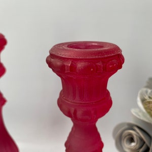 May include: A pair of red glass candle holders with a textured, ornate design. The holders are made of frosted glass and have a vintage look.
